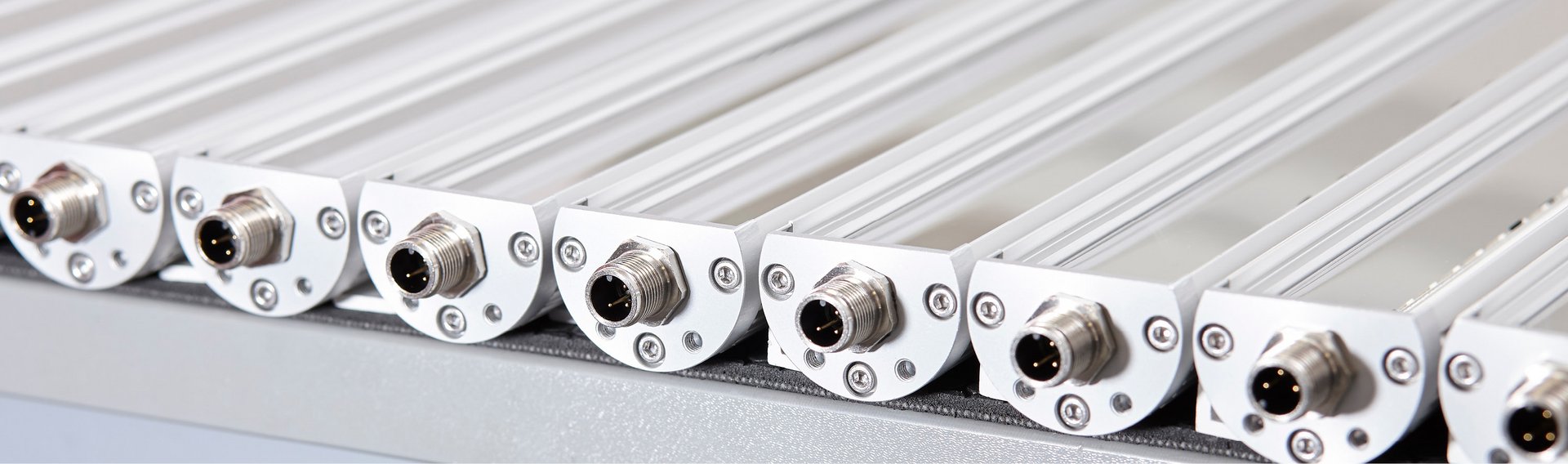 Series of industrial LED fixtures mounted closely side by side, each equipped with a metallic M12 connector on the front, installed on bright aluminum profiles.